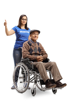 Young Woman Volunteer Pushing A Senior Man In A Wheelchair And Showing Thumbs Up