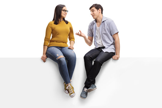 Young man sitting on a blank signboard and talking to a young woman