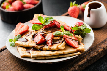 French crepes with strawberries and chocolate sauce on a plate served on wooden cutting board. Closeup view. Tasty sweet breakfast, lunch or dessert