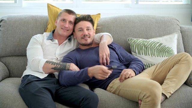 Young gay male couple relaxing on couch in living room watching tv. Caucasian, european men sitting on sofa. Using remote control. Happy gay friendship, relationship, lifestyle concept. 