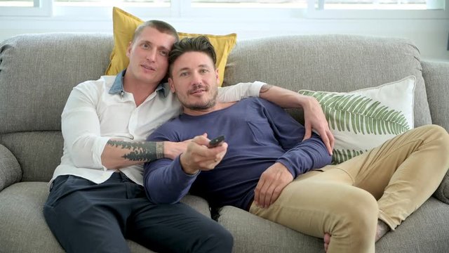 Young gay male couple relaxing on couch in living room watching tv. Caucasian, european men sitting on sofa. Trying to find the right channel. Happy gay friendship, relationship, lifestyle concept. 