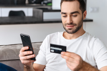 Photo of caucasian guy using smartphone and holding credit card while sitting on sofa at home
