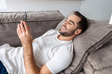 Photo of happy guy holding and using smartphone while lying on sofa in bright apartment