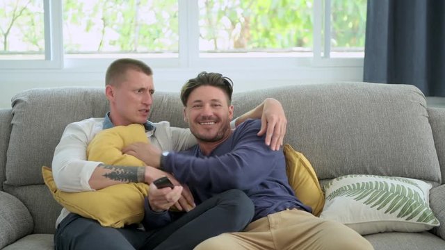 Young gay male couple relaxing on couch in living room watching tv. Caucasian, european men sitting on sofa. Teasing each other. Happy gay friendship, relationship, lifestyle concept. 