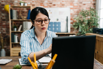 Fototapeta premium Smiling beautiful asian chinese businesswoman worker sitting at wooden desk in cozy modern office serious looking at laptop computer screen working and greenery in red brick wall. eco friendly