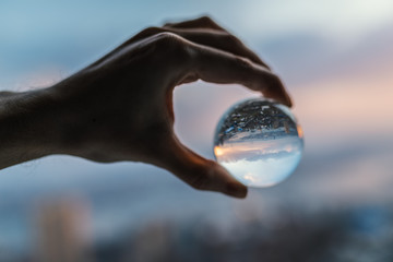 Hand holds glass ball which reflects sunset sky over city.