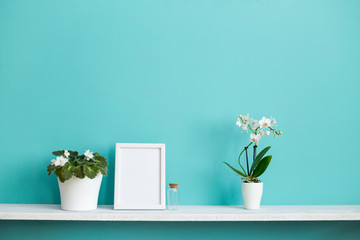 Modern room decoration with Picture frame mockup. White shelf against pastel turquoise wall with potted orchid and violet plant.