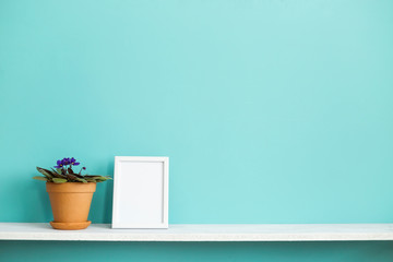 Modern room decoration with Picture frame mockup. White shelf against pastel turquoise wall with potted violet plant.