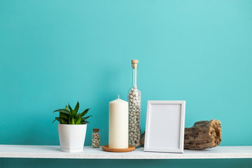 Modern room decoration with Picture frame mockup. White shelf against pastel turquoise wall with Candle and rocks in bottle. Potted snake plant
