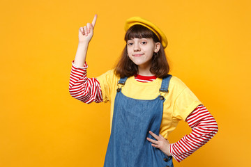 Portrait of cute girl teenager in french beret and denim sundress pointing index finger up isolated on yellow wall background in studio. People sincere emotions, lifestyle concept. Mock up copy space.