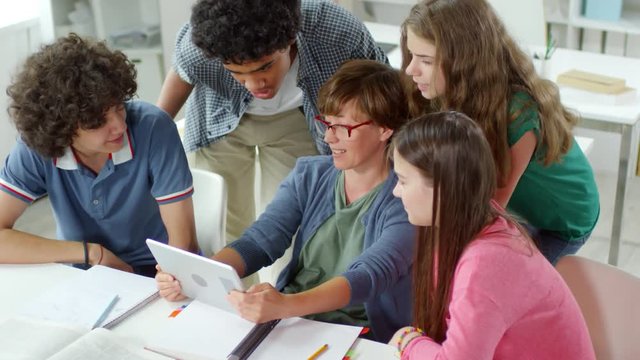 High angle view of classmates gathered around their female teacher sitting at desktop with tablet in her hands and solving test together
