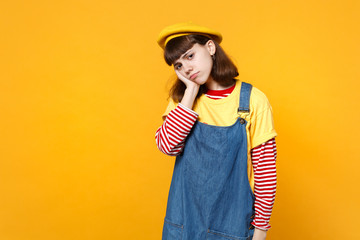 Portrait of tired dissatisfied girl teenager in french beret, denim sundress putting hand on cheek isolated on yellow wall background. People sincere emotions, lifestyle concept. Mock up copy space.