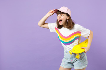 Naklejka premium Excited teen girl in vivid clothes looking aside, holding yellow skateboard, keeping mouth open isolated on violet pastel background. People sincere emotions, lifestyle concept. Mock up copy space.
