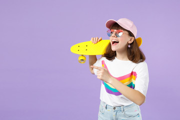 Excited teen girl in vivid clothes, eyeglasses hold yellow skateboard, pointing index finger aside isolated on violet pastel background. People sincere emotions, lifestyle concept. Mock up copy space.