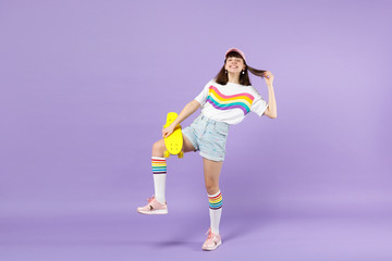 Portrait of laughing teen girl in vivid clothes standing, holding yellow skateboard isolated on violet pastel wall background in studio. People sincere emotions, lifestyle concept. Mock up copy space.