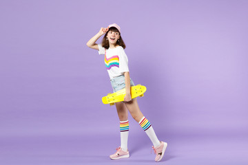 Fototapeta na wymiar Side view of cheerful teen girl in vivid clothes holding yellow skateboard, looking back isolated on violet pastel background in studio. People sincere emotions, lifestyle concept. Mock up copy space.