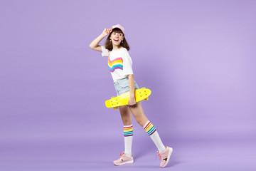 Side view of cheerful teen girl in vivid clothes holding yellow skateboard, looking back isolated on violet pastel background in studio. People sincere emotions, lifestyle concept. Mock up copy space.