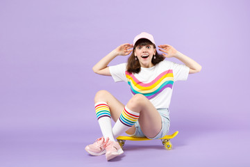 Shocked teen girl in vivid clothes sitting on yellow skateboard, keeping mouth open, rising hands isolated on violet pastel background. People sincere emotions, lifestyle concept. Mock up copy space.