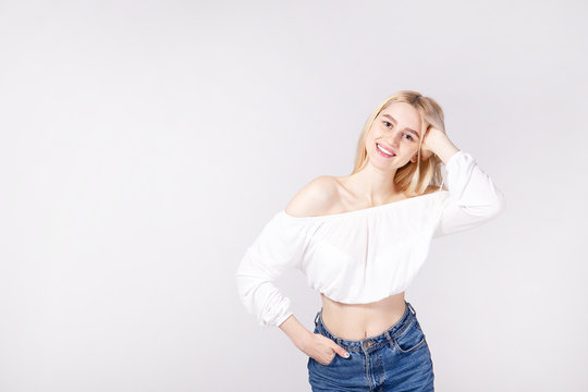 Studio Shot Of Gorgeous Young Blonde Woman With Straight Hair Wearing Off Shoulder Crop Top Sweater And High Waisted Denim Shorts. Gray Isolated Background, Copy Space, Close Up.