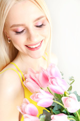 Studio portrait of gorgeous young blonde woman with long straight hair wearing bright yellow dress and holding bouquet of many tender pink tulips. Gray isolated background, copy space, close up.