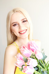 Studio portrait of gorgeous young blonde woman with long straight hair wearing bright yellow dress and holding bouquet of many tender pink tulips. Gray isolated background, copy space, close up.