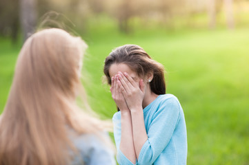 little girl in blue blouse hiding her face with both hands from another small girl outdoor
