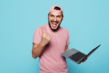 Joyful stylish student wearing pink baseball cap standing and holding laptop, fist up in success on...
