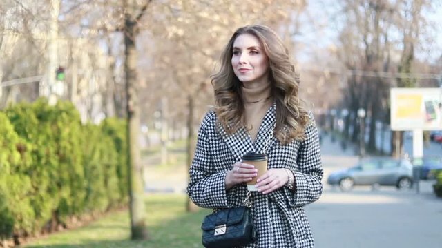 smiling elegant woman in trench coat walking and drinking coffee from paper cup . smiling woman with curly hair look at camera smile walking slow motion face sunset beautiful lady outdoor closeup cute