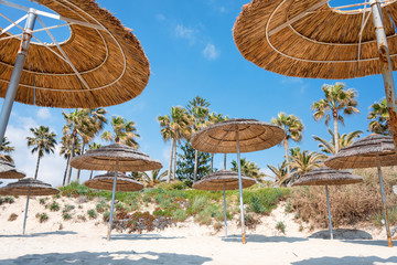 Fototapeta na wymiar Reed beach umbrellas, sunshades against blue sky on the beach. Bamboo parasols, straw umbrellas on on white sandy tropical seashore. Tropical sea beach coastline, summer holiday.