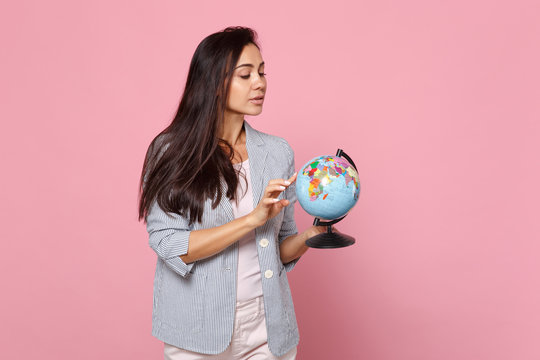 Curious young woman in striped jacket holding pointing index finger on Earth world globe isolated on pink pastel background in studio. People sincere emotions, lifestyle concept. Mock up copy space.