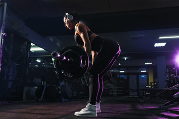 Fototapeta na wymiar Attractive fit sexy woman in wireless headphones listen music and doing crouches with a barbell in the gym. Woman training back.