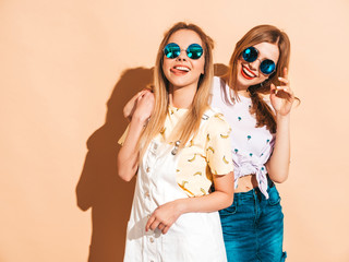 Two young beautiful smiling blond hipster girls in trendy summer colorful T-shirt clothes. Sexy carefree women posing near beige wall in round sunglasses. Positive models having fun