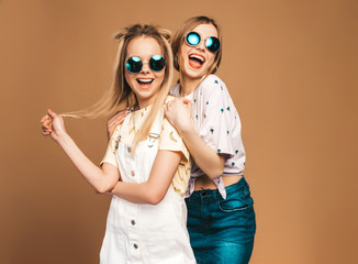 Two young beautiful smiling blond hipster girls in trendy summer colorful T-shirt clothes. Sexy carefree women posing near beige wall in round sunglasses. Positive models having fun