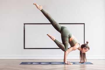 Slim charming young girl gymnast doing a complex handstand on the mat on the floor in a gymnastic suit. Concept of yoga and acrobatics. Advertising space
