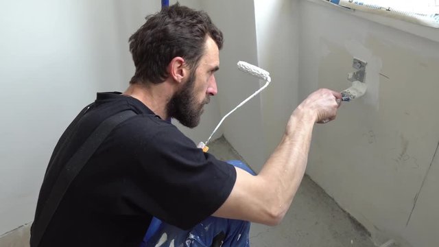 Young Caucasian Brown Haired Man with Beard and Black T-Shirt Blue Dungarees Working Trousers is Crouching Down Painting with Paint Brush and Fresh White Color the Bottom Wall Below the Window