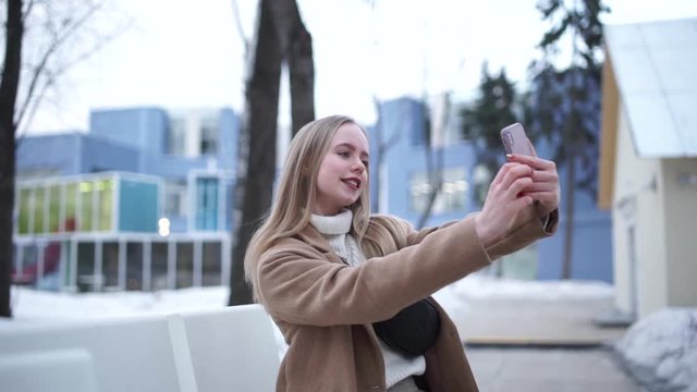 Close up of the pretty young blonde girl taking selfie photos on the smartphone device camera and posing to it. Outdoor.