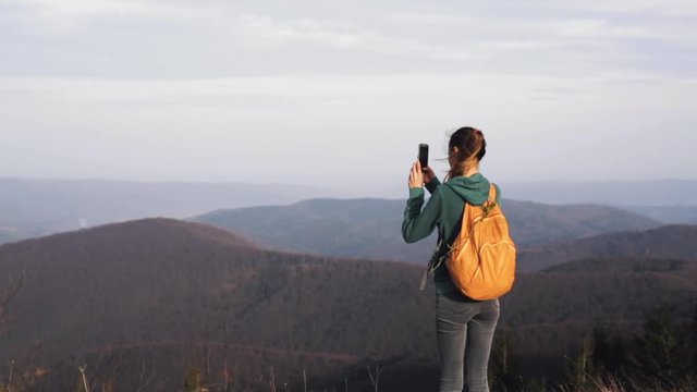 pretty woman hiker with small orange backpack is standing on edge of mountain ridge. young woman standing on mountain ridge and enjoy the view at sunset and taking a photo using phone. strong wind is