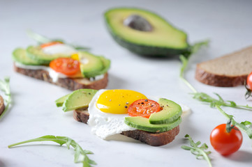 Toasts with avocado, cherry tomato and fried egg. The composition is supplemented with arugula greens. Light background. 