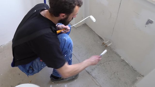 Young Caucasian Brown Haired Man with Beard and Black T-Shirt Blue Dungarees Trousers is Crouching Down Dipping in Fresh White Color Bucket with Paint Brush and Painting Bottom Wall Below the Window
