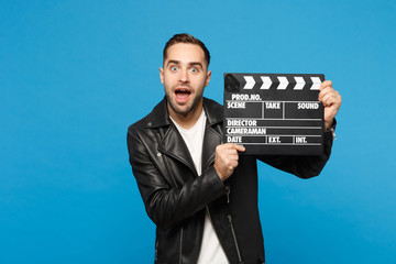 Handsome stylish young unshaven man in black jacket white t-shirt hold in hand film making clapperboard isolated on blue wall background studio portrait. People lifestyle concept. Mock up copy space.