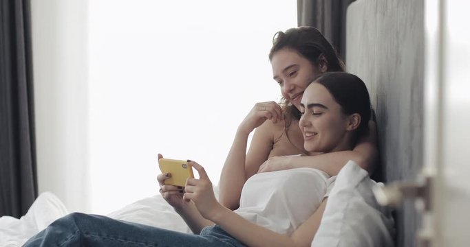 Happy lesbian couple lying in the bed hug and watch film on smart phone device. LGBT lesbian couple together. Slow motion.