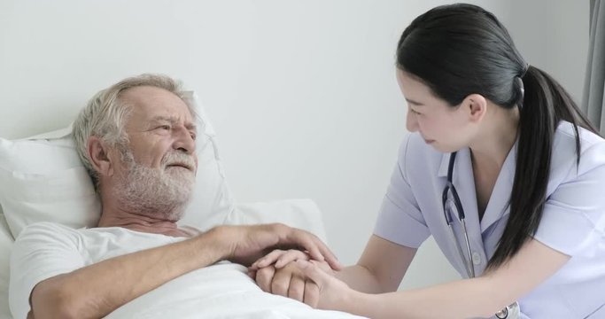 Nurse with senior patient in bedroom. Asian female nurse with bearded white male in bed. Holding hands and thumbs up. Senior lifestyle concept.