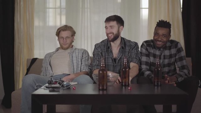 Three young men friends sitting at house on the cauch, looking tv, drinking beer with chips, watching football match and very unset and disappointed with play