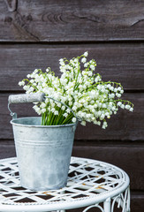 Bouquet of white cut flowers Lily of the valley (Convallaria majalis) in a zinc bucket shaped vase, outdoors on a white   fancy flower stand, brown wooden background.