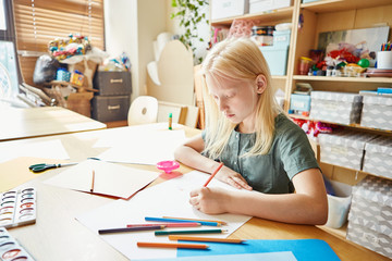 Adorable albino girl drawing on white paper sheet with colored pencils while sitting at table in...