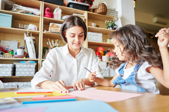 Pretty young woman smiling while teaching cute girl to draw during art class in school