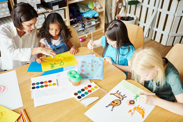 From above shot of young teacher sitting at table near little girls and helping them to paint...