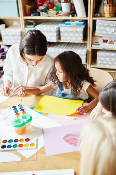 Cute girl preparing to paint while sitting near young teacher during art class in school