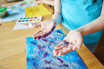 From above shot of anonymous little girl standing near table and showing hands with glitter during art class in school