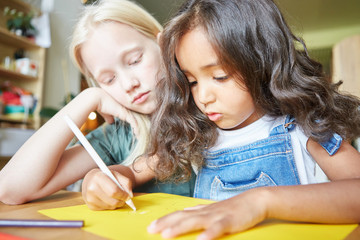 Two charming little girls sitting at desk and drawing picture on yellow paper while studying in art...